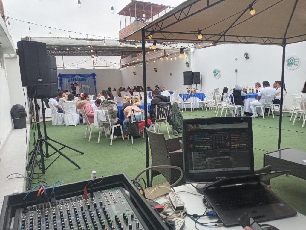 Evento al aire libre: consola de mezcla, laptop y monitor con software DJ, parlante en trípode e invitados en jardín con césped.